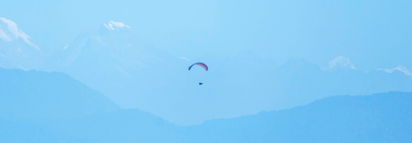 Paraglider flying high against a pale blue sky with snow-capped mountains in the background.