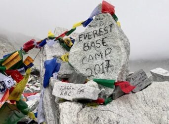 A pile of large rocks, with "EVEREST BASE CAMP 2017" painted on one, is covered in colorful prayer flags.