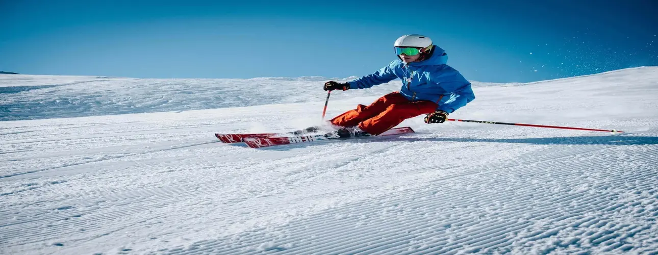 Skier in a blue jacket and orange pants carving down a groomed slope under a bright blue sky.