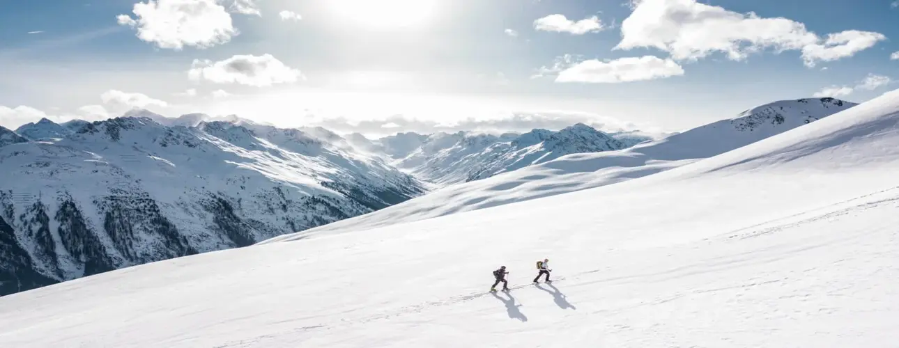 Two hikers climbing a vast, sunny, snow-covered mountain slope with a range of peaks in the background.