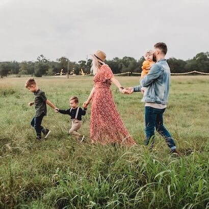 A family of five, including a woman in a long red dress and a man carrying a baby, holds hands while walking through a grassy field.