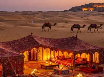 Ornate, lit desert camp and camels walking towards a distant fort across sand dunes at twilight in India.