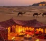Ornate, lit desert camp and camels walking towards a distant fort across sand dunes at twilight in India.
