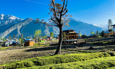 A sunlit village with wooden houses nestled in a valley beneath towering snow-capped mountains and a vibrant blue sky.