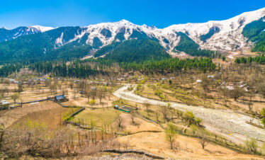 A winding river flows through a rural mountain valley village beneath snow-capped peaks and a bright blue sky.