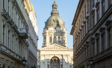 A low-angle view of St. Stephen's Basilica in Budapest, framed by the neoclassical architecture of a narrow city street.