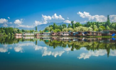A serene lake with houseboats and trees reflecting on calm water, with mountains and a clear blue sky in the background.