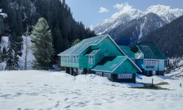 A snow-covered mountain lodge surrounded by pine trees with snow-capped peaks under a clear blue sky.