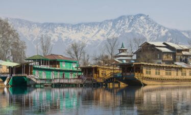 Traditional houseboats on a calm lake with snow-covered mountains and bare trees in the background under a clear sky.