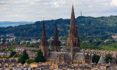 The three gothic spires of St. Mary's Episcopal Cathedral rise prominently above the historic rooftops and lush green hills of Edinburgh, Scotland.
