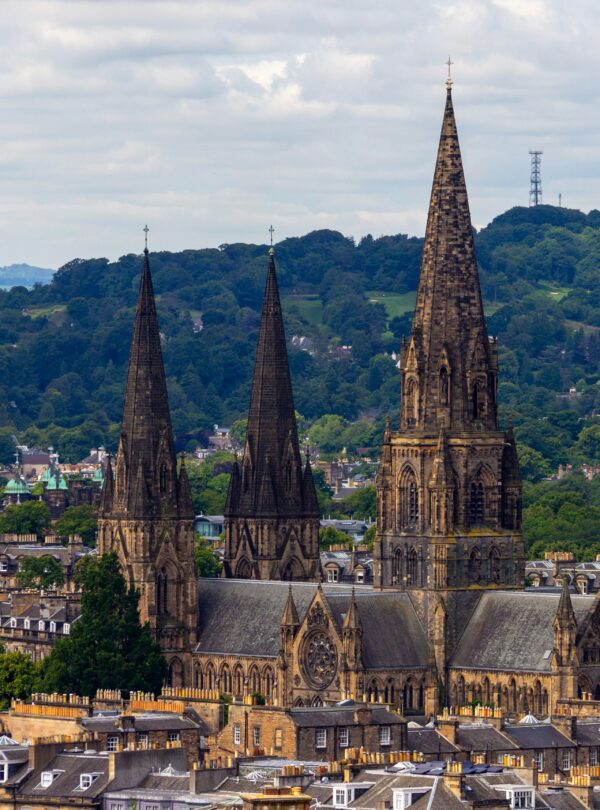 The three gothic spires of St. Mary's Episcopal Cathedral rise prominently above the historic rooftops and lush green hills of Edinburgh, Scotland.