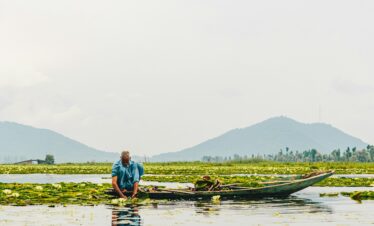 A man sitting in a small wooden boat on a lake covered with water plants, with distant hills under a hazy sky.