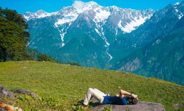A person lies on a rock in a grassy meadow overlooking a massive, snow-capped mountain range under a clear blue sky.