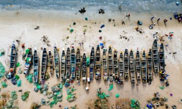 An overhead aerial view of numerous boats lined up along a sandy shoreline with scattered nets and fishing gear.