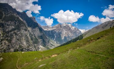 A scenic mountain valley with green meadows and rugged cliffs leading to snow-capped peaks under a bright blue sky with clouds.
