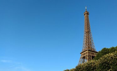 The Eiffel Tower rises above lush green treetops into a clear blue sky.