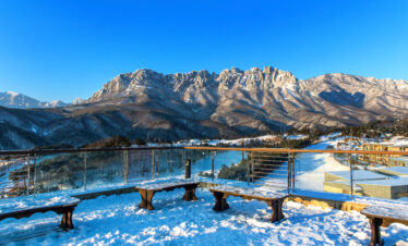 A snow-covered observation deck with wooden benches overlooks a dramatic, jagged mountain range under a clear blue sky.