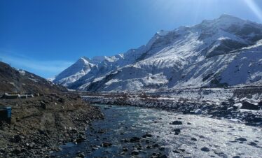 A rocky river flows through a high-altitude valley toward towering snow-covered mountains under a bright sun.