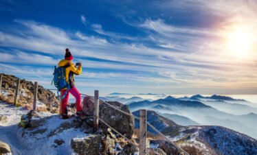A hiker in a yellow jacket takes a photo of a breathtaking mountain range emerging from a sea of clouds under a bright, sunlit sky.